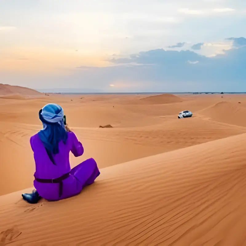 Traveler in blue robe taking photos of the vast Erg Chebbi dunes with a private 4x4 in the distance on the Marrakech to Merzouga Desert Tour