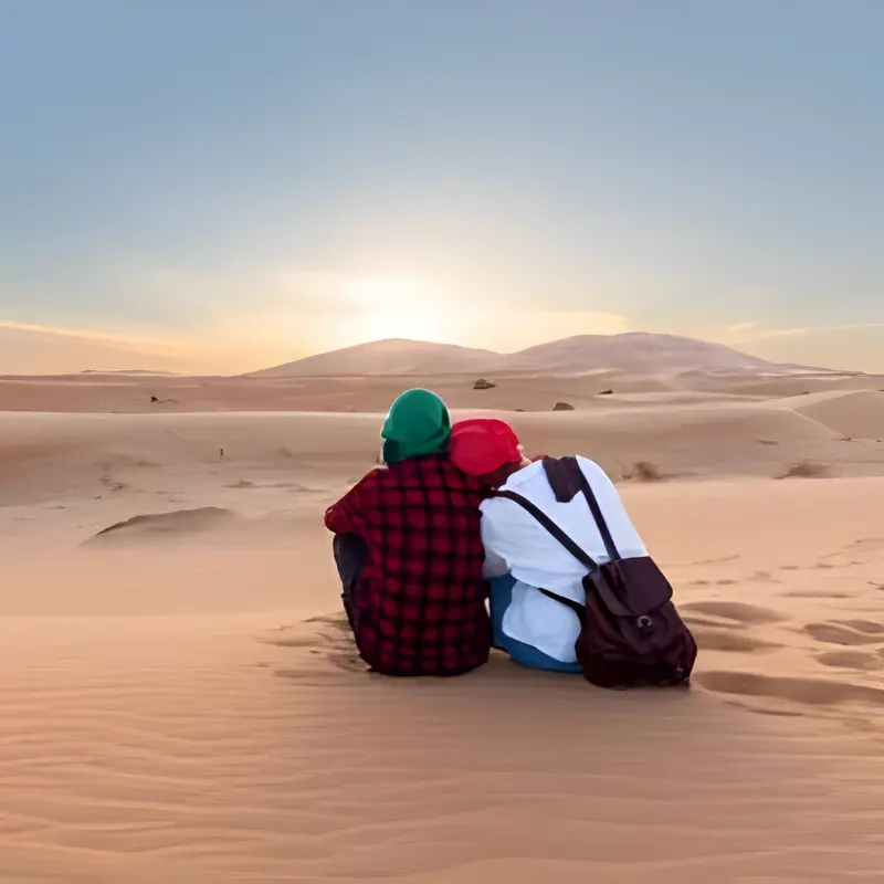 Two travelers sitting together watching sunrise over the Erg Chebbi dunes on a 4-Day Marrakech to Merzouga Desert Tour